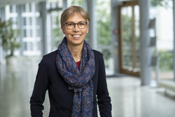 KI generiert: Auf dem Bild ist eine lächelnde Person in einem Innenraum zu sehen, die eine Brille und einen gemusterten Schal trägt. Der Hintergrund zeigt einen hellen, modernen Korridor.