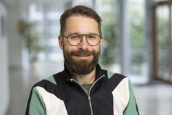KI generiert: Das Bild zeigt einen lächelnden Mann mit Brille und Bart, der in einem hellen Raum steht. Der Fokus liegt auf seinem Gesicht und seiner entspannten Ausstrahlung.
