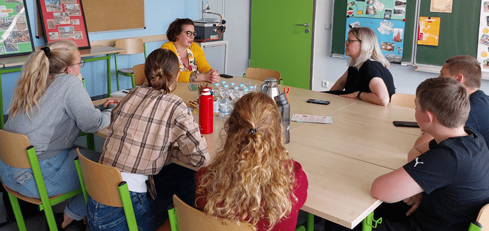 KI generiert: Eine Gruppe von Menschen sitzt in einem Klassenzimmer um einen Tisch und diskutiert. Es scheint sich um ein Gespräch oder eine Besprechung zu handeln.