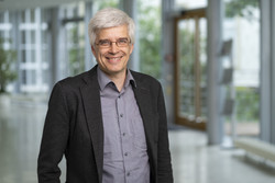 KI generiert: Das Bild zeigt einen Mann mittleren Alters mit grauem Haar und Brille, der lächelnd in einem modernen, lichtdurchfluteten Innenraum steht. Er trägt ein dunkles Sakko und ein graues Hemd.