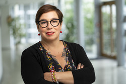KI generiert: Das Bild zeigt eine Person mit Brille und dunklen Haaren, die vor einer verschwommenen Innenraumszene mit verschränkten Armen steht. Die Person trägt einen blumenbedruckten Oberteil unter einem schwarzen Cardigan.