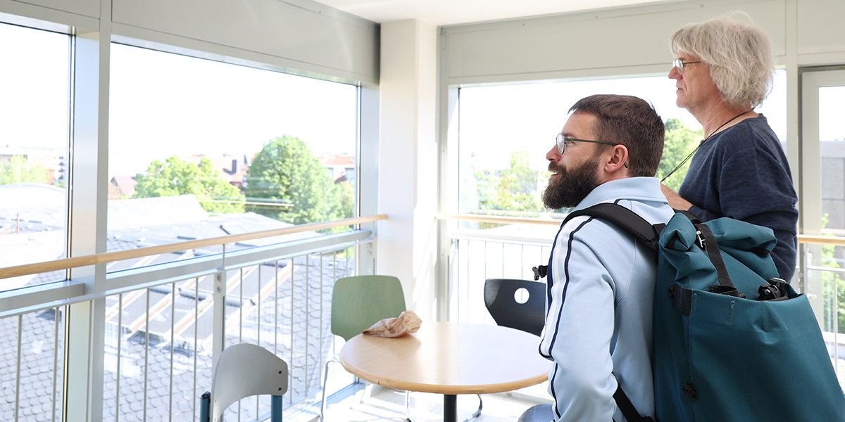 KI generiert: Zwei Personen stehen in einem hellen Raum vor großen Fenstern und schauen nach draußen. Im Vordergrund ist ein runder Tisch mit Stühlen zu sehen.