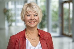 KI generiert: Das Bild zeigt eine lächelnde Person mit blonden Haaren in einem roten Jackett, in einem hellen, unscharfen Innenraum. Der Fokus liegt auf dem freundlichen Ausdruck der Person.