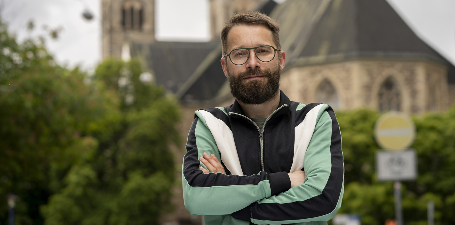KI generiert: Das Bild zeigt eine Person mit Brille und Bart, die vor einer alten Kirche steht. Die Person hat die Arme verschränkt und trägt eine zweifarbige Jacke.