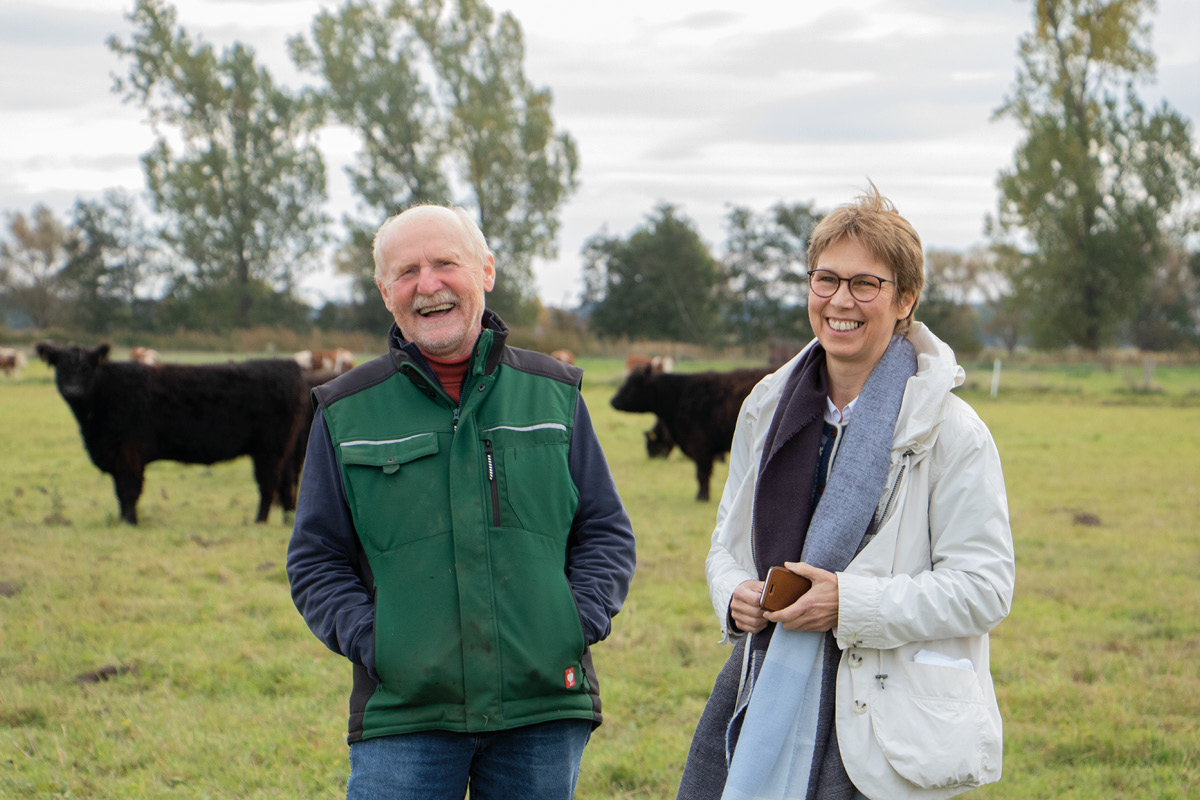Dorothea Frederking gemeinsam mit einem Landwirt auf einer Weide. Im Hintergrund sieht man Kühe und Weidetiere.