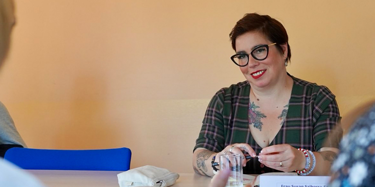 KI generiert: Eine Person sitzt an einem Tisch in einem Gespräch mit anderen. Sie trägt Brille, hat tätowierte Arme und trägt ein kariertes Oberteil.