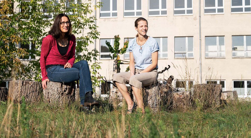 Katharina Bensch und Stefanie Wischer sitzen auf Holzscheiten vor einem Gebäude, vor dem eine Wiese liegt. 