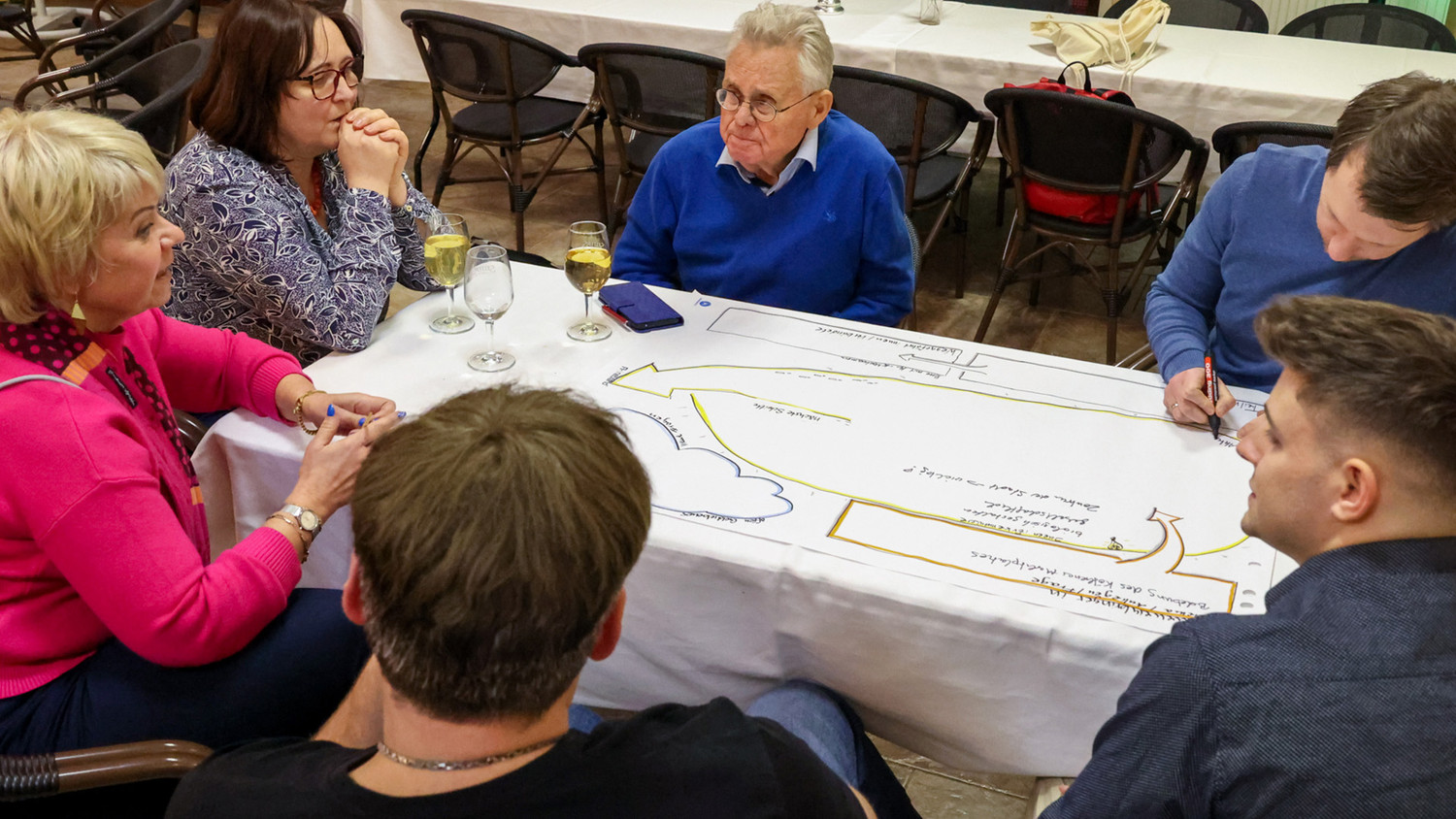KI generiert: Eine Gruppe von sechs Personen sitzt um einen Tisch und diskutiert oder arbeitet an einem Diagramm auf einem großen Blatt Papier. Die Szene scheint eine kollaborative Besprechung oder ein Workshop zu sein, bei dem Details notiert und geteilt werden.