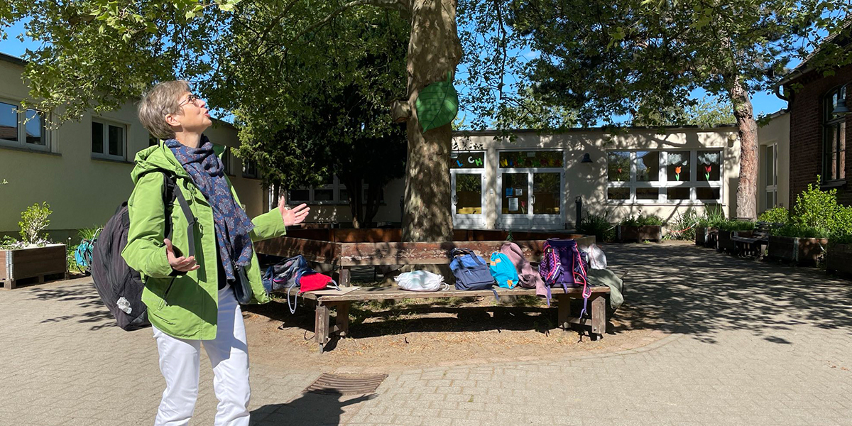 KI generiert: Eine Person steht in einem Schulhof und betrachtet die Umgebung, während mehrere Taschen auf einer Bank liegen. Im Hintergrund ist ein Schulgebäude zu sehen.