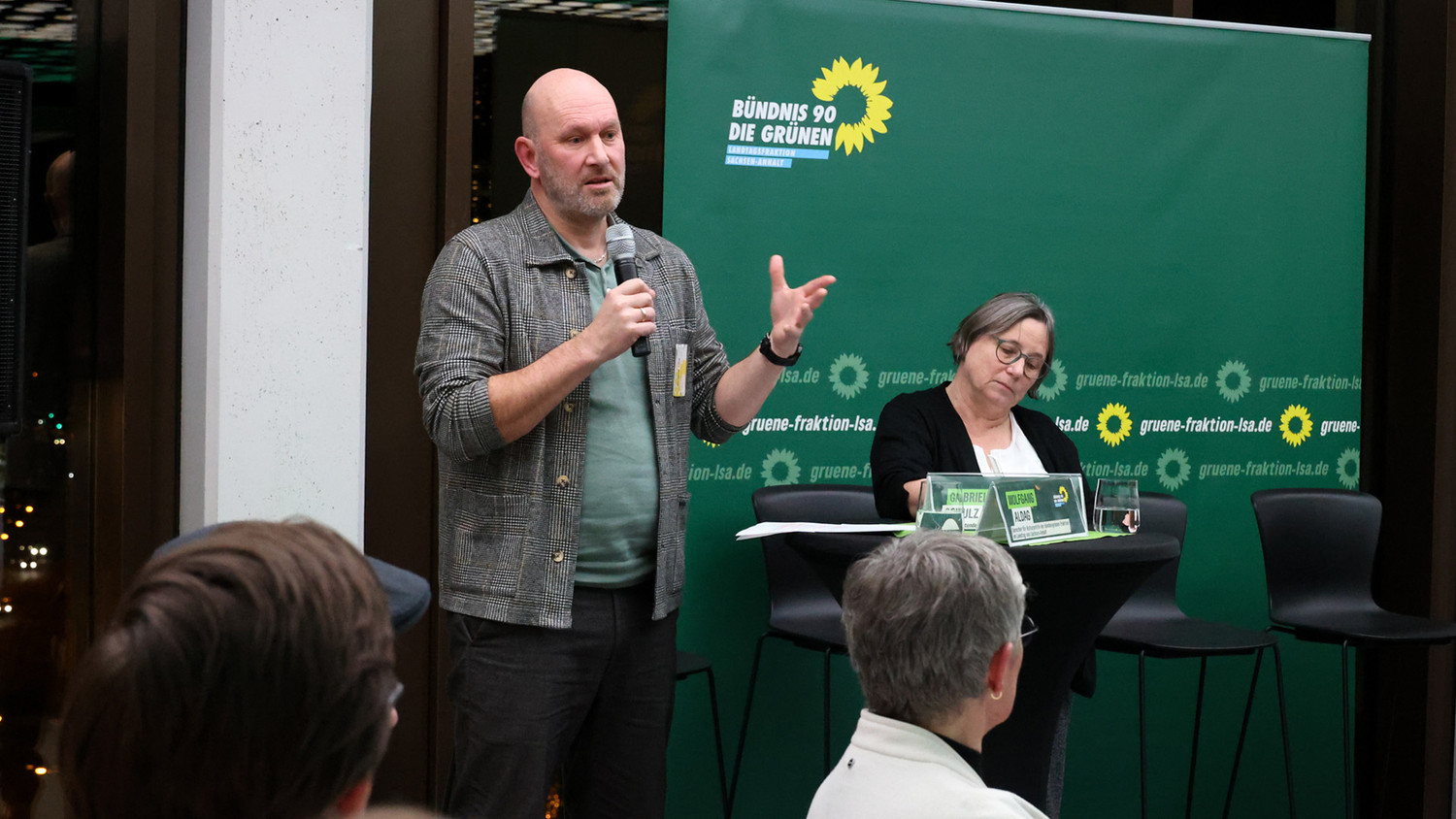 KI generiert: Auf dem Bild spricht ein Mann mit einem Mikrofon vor einem Banner von Bündnis 90/Die Grünen, während eine Frau daneben steht und Notizen macht. Der Hauptinhalt des Bildes ist eine Präsentation oder Diskussion im Rahmen einer Veranstaltung der Partei.