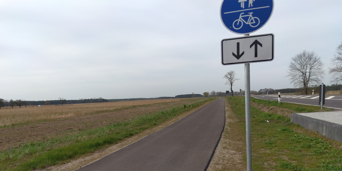 KI generiert: Das Bild zeigt einen kombinierten Fußgänger- und Radweg in einer ländlichen Umgebung mit entsprechender Beschilderung. Der Weg verläuft parallel zu einem Feld und einer Straße.