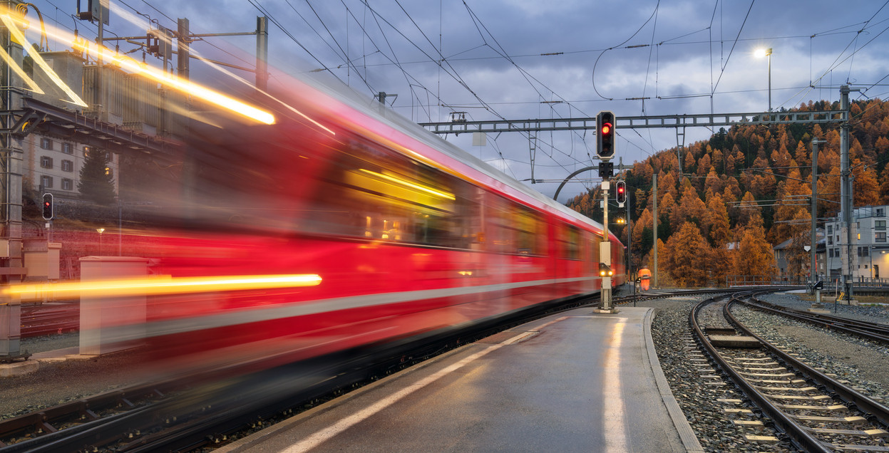 KI generiert: Das Bild zeigt einen roten Zug in Bewegung auf einem Bahnhofsgleis, was durch die Bewegungsunschärfe hervorgehoben wird. Im Hintergrund sind herbstliche Bäume und städtische Gebäude zu sehen.