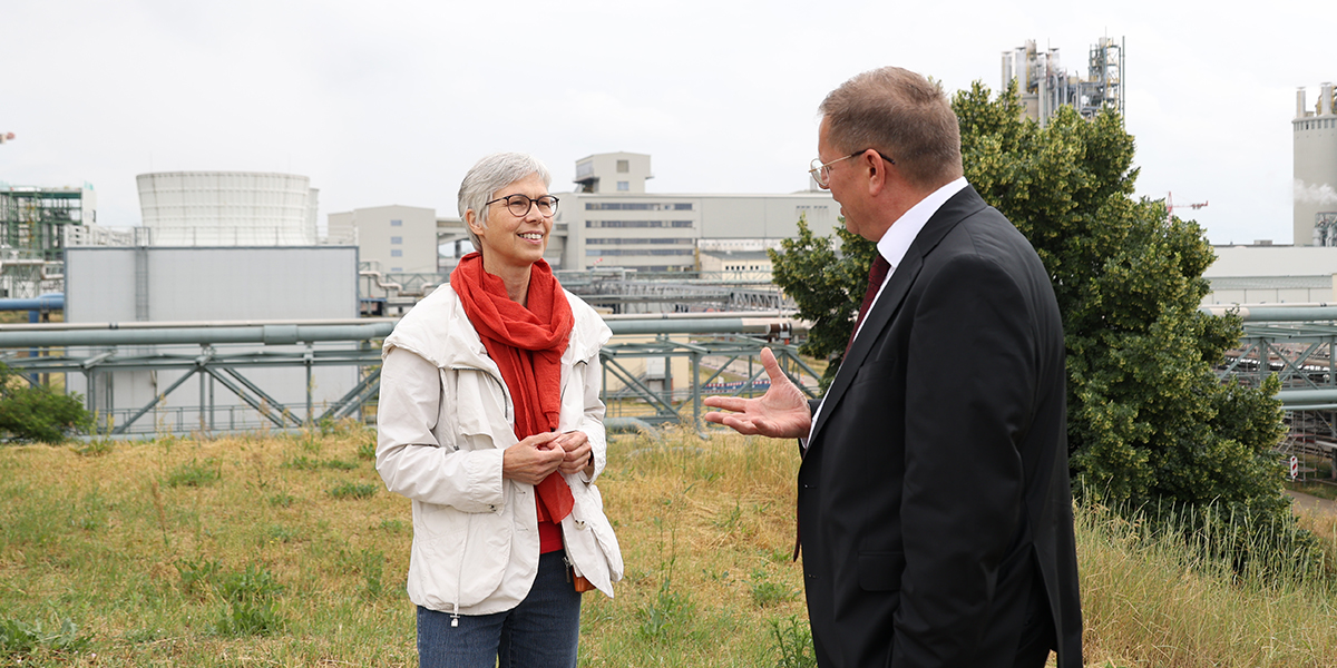 KI generiert: Zwei Personen unterhalten sich freundlich vor einer industriellen Kulisse mit Gebäuden und Rohrleitungen. Im Vordergrund steht eine Wiese, und im Hintergrund sind mehrere Fabrikstrukturen sichtbar.