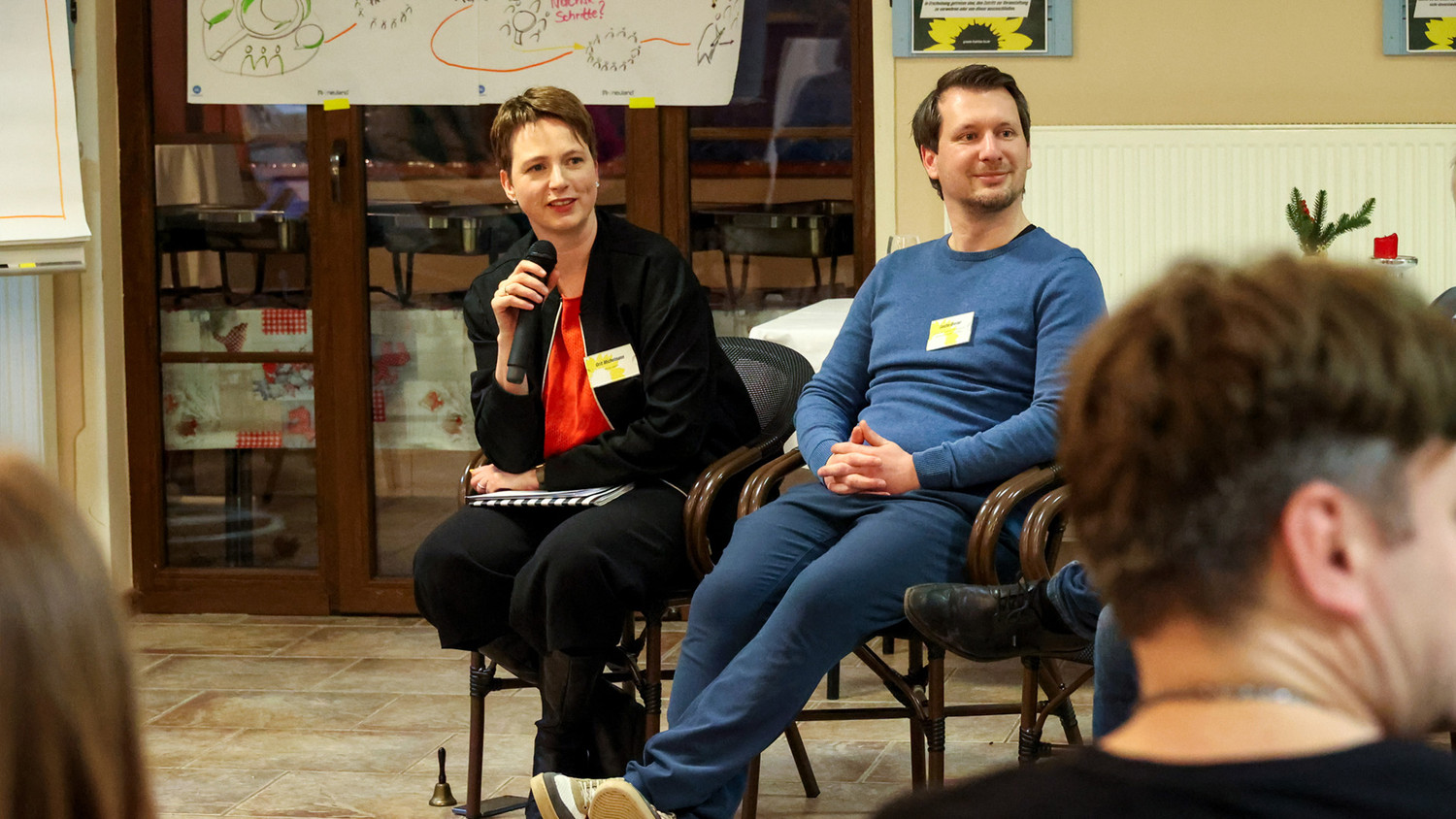 KI generiert: Das Bild zeigt eine Frau und einen Mann, die an einer Diskussionsveranstaltung teilnehmen. Die Frau hält ein Mikrofon und spricht, während der Mann aufmerksam zuhört.