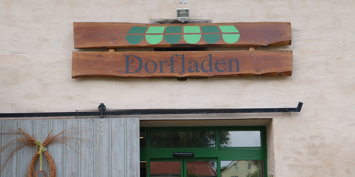 KI generiert: Das Bild zeigt ein Holzschild mit der Aufschrift "Dorfladen" über einem Eingang. Der Eingang ist durch ein Fenster mit einem grünen Rahmen und einer Jalousie aus Holz gekennzeichnet.