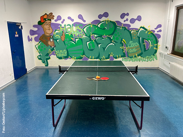 Eine Tischtennisplatte vor einer mit Graffiti besprühten Wand, auf der Tischtennisball und Schläger liegen.