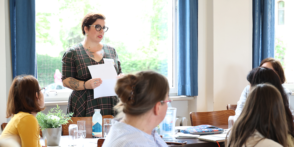 KI generiert: Das Bild zeigt eine Frau, die in einem Raum vor mehreren Personen steht und ein Dokument hält, als ob sie eine Präsentation oder Rede hält. Die Anwesenden sitzen an einem Tisch, der mit Getränken und Unterlagen bedeckt ist.