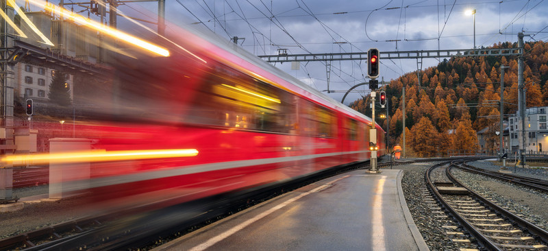 KI generiert: Das Bild zeigt einen roten Zug, der mit hoher Geschwindigkeit durch einen Bahnhof fährt und eine Bewegungsspanne aufweist. Im Hintergrund sind herbstlich gefärbte Bäume und ein bewölkter Himmel zu sehen.