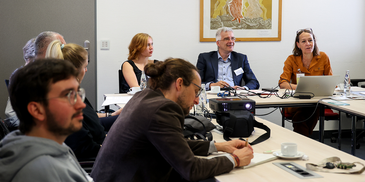 KI generiert: Das Bild zeigt eine Gruppe von Menschen, die in einem Konferenzraum in einer Besprechung oder Diskussion versammelt sind. Auf dem Tisch stehen Arbeitsmaterialien und ein Projektor ist in Betrieb.