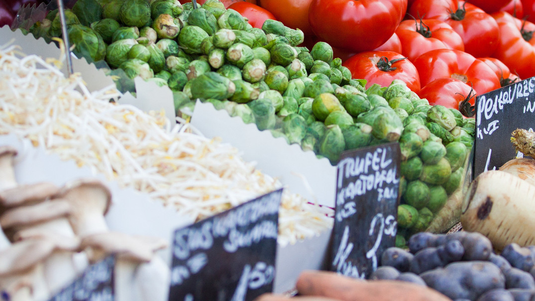 KI generiert: Das Bild zeigt einen Marktstand mit frischem Gemüse und Obst, darunter Rosenkohl, Tomaten und Pilze. Verschiedene handgeschriebene Preisschilder sind sichtbar.