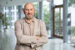 KI generiert: Das Bild zeigt einen lächelnden Mann in einem hellen Anzug, der mit verschränkten Armen in einem modernen, lichtdurchfluteten Raum steht. Der Hintergrund deutet auf ein Bürogebäude oder eine ähnliche Einrichtung hin.